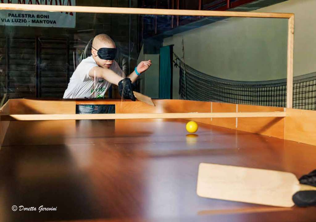  SHOWDOWN SPORT PRATICATO DAI DISABILI VISIVI – UNA SORTA DI PING PONG CON PALLINA SONORA E PALETTA DI LEGNO I CUI GIOCATORI BENDATI SI SFIDANO ALL’INTERNO DI UN TAVOLO CON PORTE E SPONDE.  NELLA FOTO: ALLA PALESTRA BONI IN PRIMO PIANO UN GIOVANE ATLETA CON MAGLIETTA BIANCA BENDATO CON APPOSITA MASCHERINA NERA IN RISPOSTA AL LANCIO DELL’AVVERSARIO 
ALL’INTERNO DEL TAVOLO DI LEGNO CON SPONDE PERIMETRICHE DI CONTENIMENTO SUDDIVISO A METÀ CAMPO DA UN PANNELLO IN PLEXIGLASS. NELL’ALTRA METÀ CAMPO IN PRIMO PIANO LA PALETTA RETTANGOLARE IMPUGNATA DALL’AVVERSARIO.