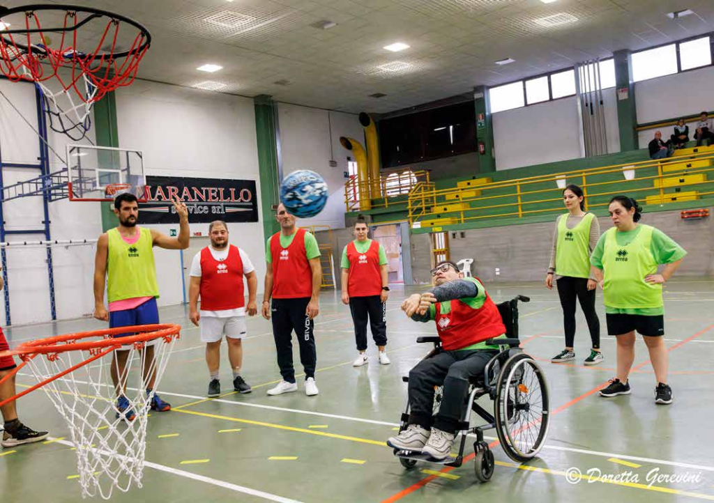 IL BASKET È INTEGRATO QUANDO PRATICATO ANCHE DALLE PERSONE CON DISABILITÀ MOTORIA: NELLA FOTO IN PALESTRA DUE SQUADRE DI GIOVANI ATLETI CON PETTORINE ROSSE E GIALLE SI SFIDANO. IN PRIMO PIANO UNO DI LORO IN CARROZZINA CON PETTORINA ROSSA IN FASE DI LANCIO CON LA PALLA A MEZZ’ARIA VERSO IL CANESTRO