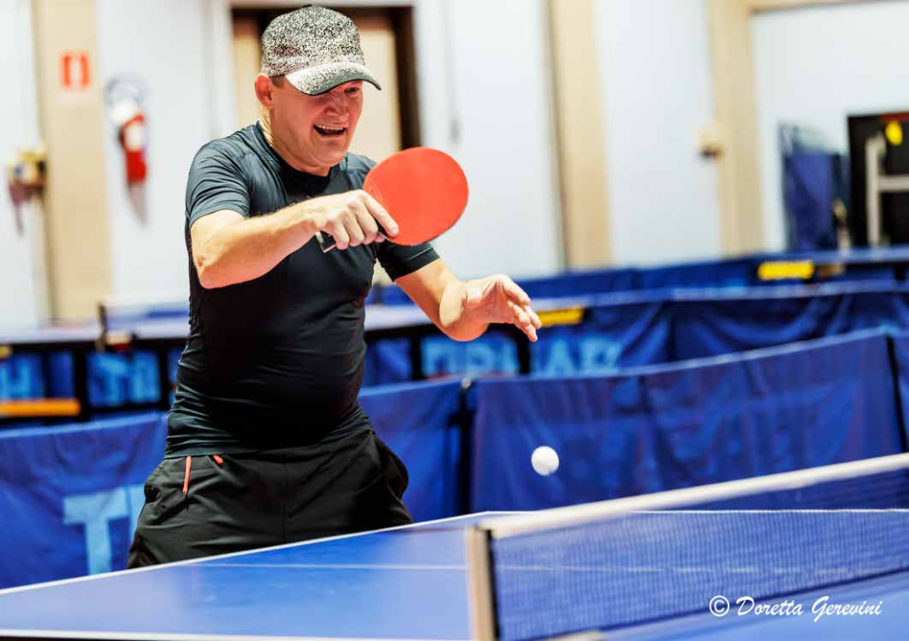 TENNIS TAVOLO: NELLA FOTO IN PALESTRA UN GIOCATORE CON TUTA NERA, CAPPELLINO NERO E BIANCO IMPUGNA UNA RACCHETTA ROSSA NELL’ATTO DI RECUPERARE LA PALLINA RIMBALZATA SUL TAVOLO DA PING PONG