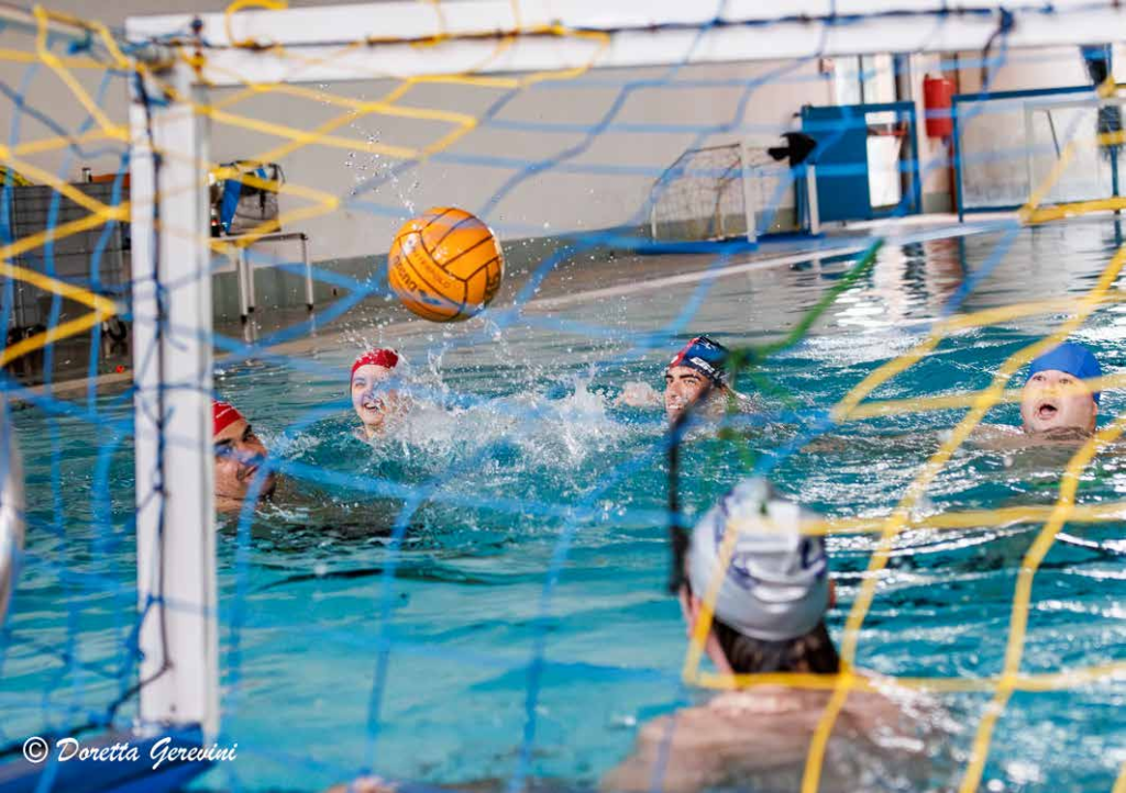 PALLANUOTO: NELLA FOTO IN UNA PISCINA CINQUE GIOCATORI IMMERSI IN ACQUA AZZURRA CON CUFFIE COLORATE E LA PALLA ARANCIONE A MEZZ’ARIA INTENTI A FARE GOAL NELLA PORTA DIFESA DAL PORTIERE DELLA SQUADRA AVVERSARIA