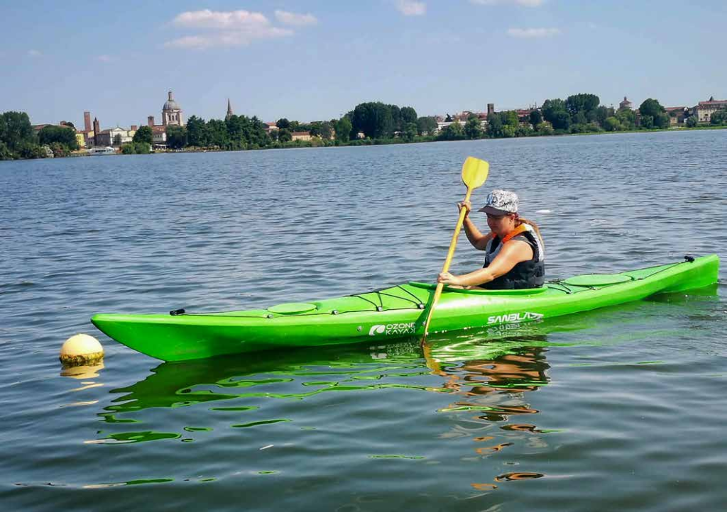 CANOA: NELLA FOTO NEL LAGO DI MANTOVA SU UNA CANOA VERDE, UNA RAGAZZA CHE INDOSSA GIUBBETTO DI SALVATAGGIO E CAPPELLO BIANCO CON VISIERA NERA STA PAGAIANDO. SULLO SFONDO LA SPONDA DEL LAGO DI MEZZO CON IN EVIDENZA LA CUPOLA DI SANT’ANDREA.