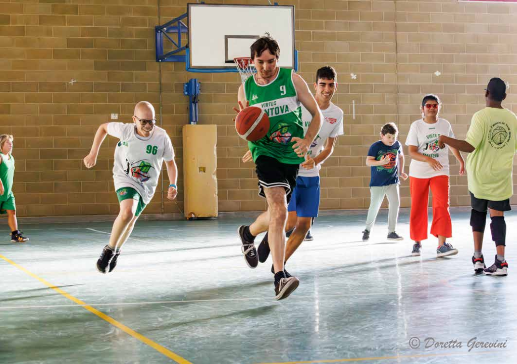 BASKET BELL LA PALLACANESTRO CON PALLA SONORA PRATICATA ANCHE DALLE PERSONE CIECHE E IPOVEDENTI: NELLA FOTO IN PALESTRA SEI RAGAZZI IN TENUTA SPORTIVA NELL’ATTO DI INSEGUIRE UN SETTIMO GIOCATORE CHE STA PALLEGGIANDO VERSO IL CANESTRO