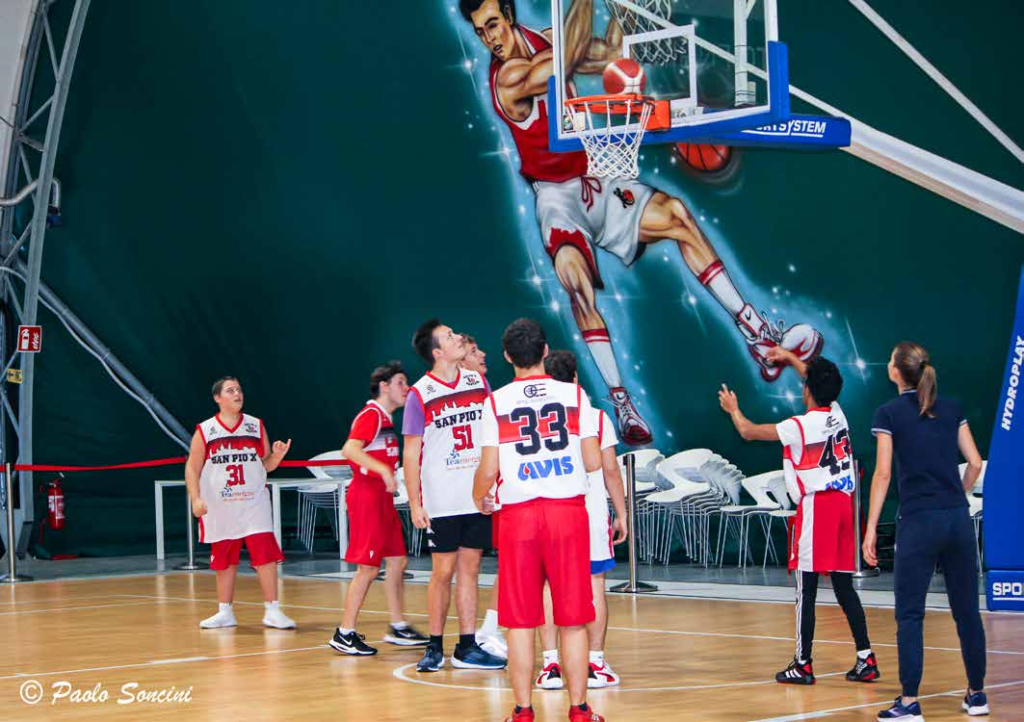 BLU BASKET: NELLA FOTO IN PALESTRA SETTE RAGAZZI IN DIVISA SPORTIVA BIANCA E ROSSA, MENTRE GUARDANO IL CANESTRO APPENA CENTRATO; SULLO SFONDO LA GIGANTOGRAFIA DI UN GIOCATORE DELL’NBA 