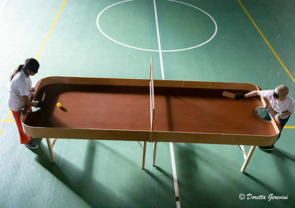 SHOWDOWN SPORT PRATICATO DAI DISABILI VISIVI – TAVOLO DA SHOWDOWN VISTO DALL’ALTO, UN BEL TAVOLO DI LEGNO CON SPONDE PERIMETRICHE DI CONTENIMENTO SUDDIVISO A METÀ CAMPO DA UN PANNELLO IN PLEXIGLASS. NELLA FOTO: DUE GIOVANI ATLETI ENTRAMBI CON MAGLIETTA BIANCA E BENDATI CON APPOSITA MASCHERINA NERA, MUNITI DI GUANTO E PALETTA SI SFIDANO FACENDO SCIVOLARE SUL TAVOLO UNA PALLINA SONORA GIALLA 